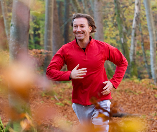 A smiling man in a red jacket jogging through a scenic forest in autumn.