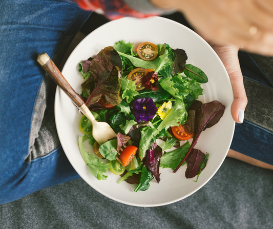 Fresh green salad with mixed greens, cherry tomatoes, and purple edible flowers in a white bowl.