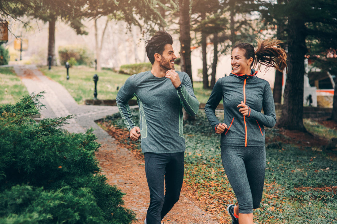 A smiling man and women jogging together along a park path lined with pine trees.