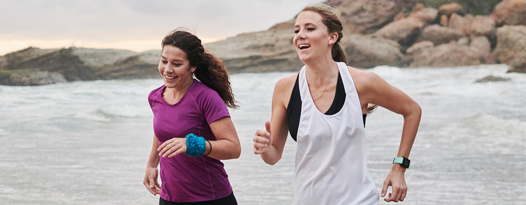 Two smiling women in athletic wear jogging together along a scenic beach shoreline.